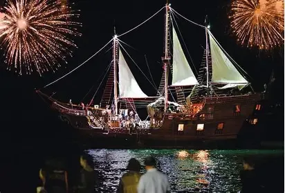 En la imagen se observa el imponente Galeón Pirata Marigalante navegando de noche por la Bahía de Banderas. El barco, totalmente iluminado con luces cálidas, destaca sobre el mar oscuro mientras el cielo nocturno se ilumina con un espectacular show de juegos pirotécnicos y fuegos artificiales de col