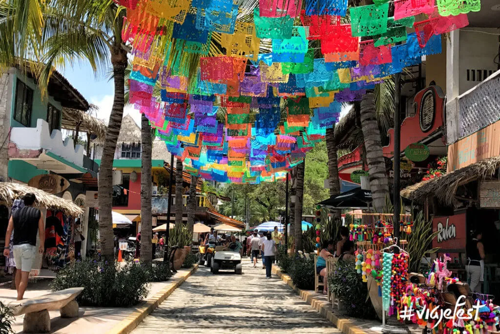 Vista de la icónica calle principal de Sayulita, rodeada de coloridas banderas decorativas, tiendas artesanales, galerías de arte, restaurantes locales y un ambiente vibrante que refleja el encanto bohemio de este famoso Pueblo Mágico de la Riviera Nayarit.